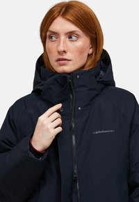 Roodharige vrouw in een zwarte Peak Performance jas met capuchon, die de rits vastmaakt en naar links kijkt.
