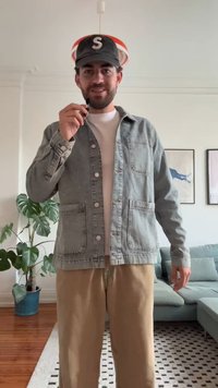 Grijze denimjas met twee voorkruisen, knoopsluiting en puntige kraag. Draag een witte t-shirt met beige broek. Zwarte pet met de letter "S."
