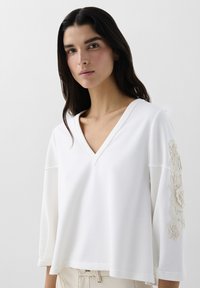 Pull blanc à col en V avec des manches trois-quarts, ornée d'une broderie florale texturée sur la manche gauche et d'une coupe décontractée. Tissu doux.