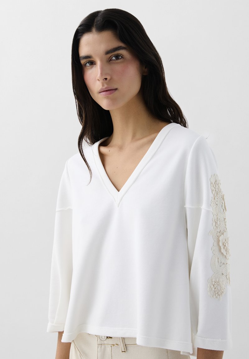 Pull blanc à col en V avec des manches trois-quarts, ornée d'une broderie florale texturée sur la manche gauche et d'une coupe décontractée. Tissu doux.
