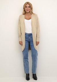 Beige pletená cardigan s texturovaným vzorem, otevřený přední díl, dlouhé rukávy a boční rozparky. Kombinováno se světlými modrými džíny a černými kotníkovými botami.