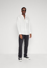 Camicia bianca a maniche lunghe in lino con taschino, abbinata a pantaloni grigio scuro e sneakers nere, accessoriate con occhiali da sole neri.