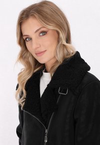 Schwarze Kunstlederjacke mit einem flauschigen schwarzen Sherpa-Kragen und Reißverschluss. Verfügt über eine seitliche Schnalle und eine glatte, matte Textur.