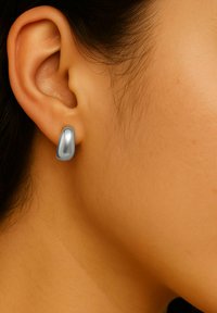 Sølvmetallisk skulpturell ørering med en jevn, blank overflate som danner en abstrakt form som omfavner øreflippen tett.