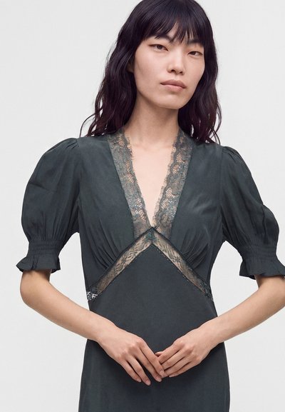 Femme aux cheveux noirs portant une robe sombre avec des manches bouffantes et une bordure en dentelle sur le décolleté en V et la taille, debout, les mains jointes.