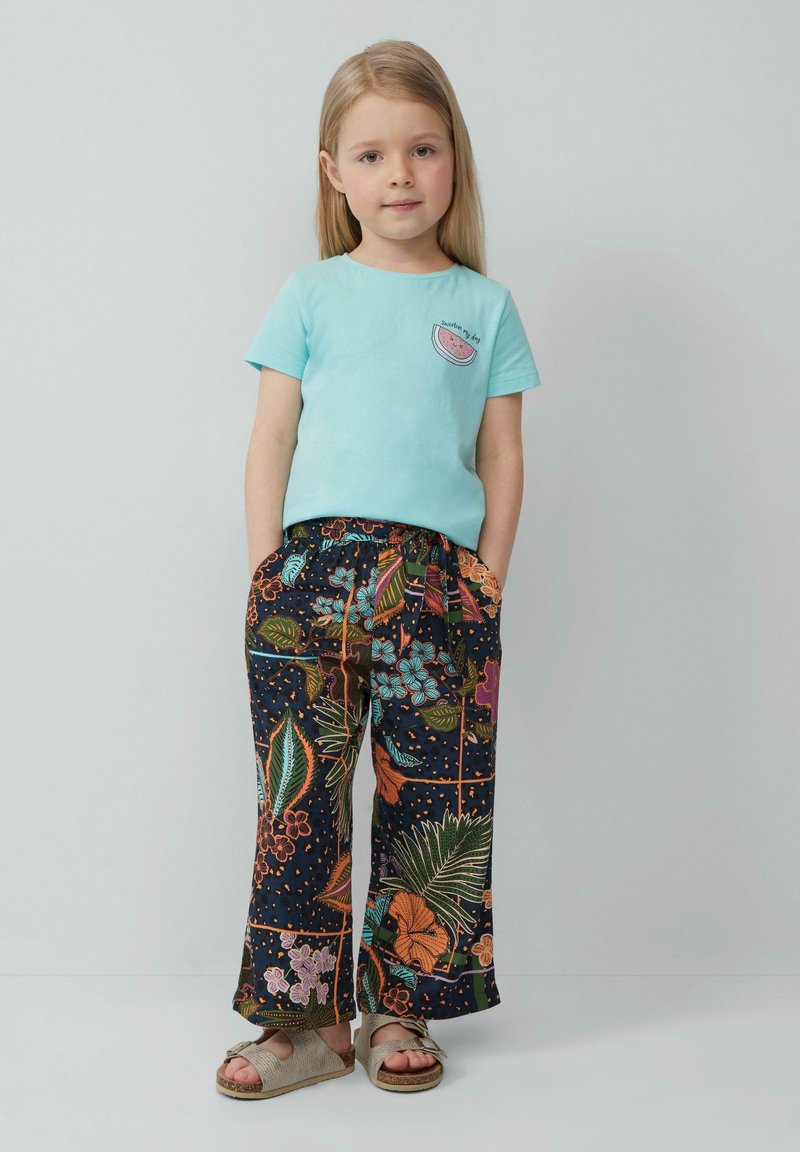 Lichtblauwe katoenen t-shirt met een watermeloenafbeelding; donkere, gebloemde broek met bloemen en bladeren; grijze sandalen met strikdetail.