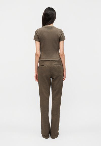 Γυναίκα με μακριά σκουρόχρωμα μαλλιά που στέκεται με την πλάτη, φορώντας ένα ταιριαστό ελιά πράσινο τοπ με κοντά μανίκια και παντελόνι με φαρδιά μπατζάκια και πίσω τσέπες.