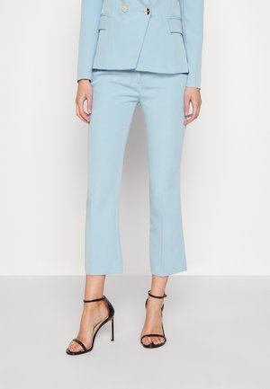 Vrouw draagt lichtblauwe op maat gemaakte broek en bijpassende blazer met zwarte hoge hak sandalen met enkelbandje, staand tegen een effen achtergrond.