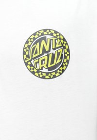 Λευκή μπλούζα που διαθέτει έναν κυκλικό γραφικό σχεδιασμό με το κείμενο "SANTA CRUZ" σε έντονο κίτρινο σε μαύρο φόντο, περιβαλλόμενο από σχέδιο με καρό μοτίβο.