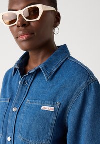 Denim srajca v srednji modri barvi z gumbnim zapiranjem, žepom na prsih in oznako Calvin Klein. Okrašena s klasičnim ovratnikom in podrobnostmi v šivanju.