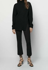 Czarny, żeberkowy sweter z wysokim kołnierzem o asymetrycznym dole, zestawiony z dopasowanymi czarnymi spodniami i czarnymi sandałami na wysokim obcasie z paskami.
