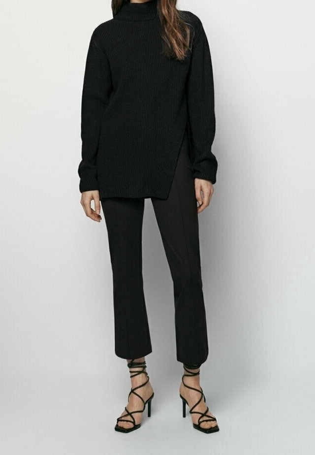 Czarny, żeberkowy sweter z wysokim kołnierzem o asymetrycznym dole, zestawiony z dopasowanymi czarnymi spodniami i czarnymi sandałami na wysokim obcasie z paskami.