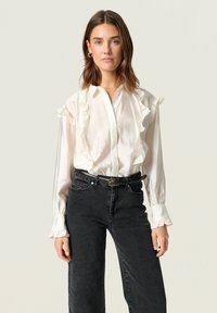 Hvidt, riflet bluse med lange ærmer, som har en knaplukning foran og blød glans, parret med højtaljede sorte denimjeans og et bælte.