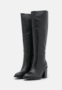 Bottes noires montantesjusqu'au genou en cuir lisse, avec un bout arrondi et un talon épais. Le design est épuré avec un minimum d'accessoires métalliques.