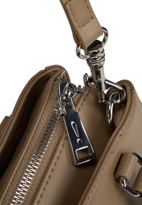 Torbica v bež barvi z gladko usnjeno teksturo, kovinsko srebrno zadrgo in kovinskimi detajli. Pogledu od blizu poudarja podrobnosti in šivanje.