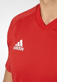 Torso masculino con una camiseta deportiva roja de Adidas con escote en V y el logo en el lado izquierdo del pecho, sobre un fondo blanco.