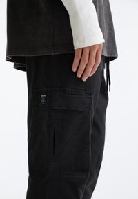 Schwarze Cargo-Hosen mit Taschen und genähten Details. Kombiniert mit einem dunklen Oberteil mit einem helleren, gerippten Ärmel. Insgesamt glatte Textur.