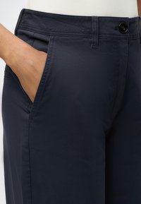 Personne portant un pantalon bleu foncé et un haut blanc, la main gauche placée dans la poche avant du pantalon.