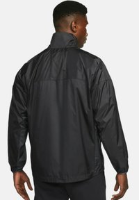 Svart, lättviktig jacka tillverkad av slätt, glänsande material. Har en hög krage och en avslappnad passform med snedställda axelsömmar och elastiska ärmslut.