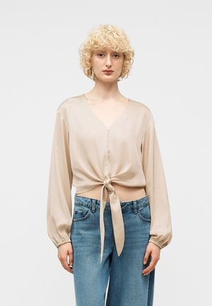 Person med krøllete blondt hår iført en beige knappet bluse knyttet i midjen og blå jeans, stående mot en ensfarget bakgrunn.