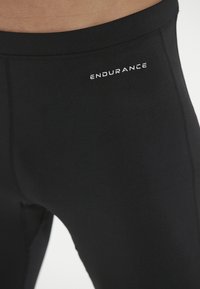 Svarta träningsleggings tillverkade av elastiskt material som har en slät yta, med en vit "ENDURANCE"-logotyp nära midjan.