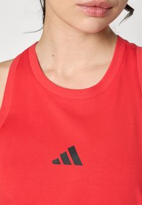 Raudonas berankovis sportinis marškinėlis, pagamintas iš lygaus audinio, su juodu Adidas logotipu priekyje, apvaliu iškirpimu ir banguotais kraštais.