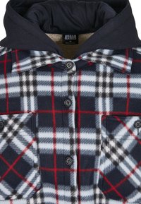 Camisa de franela a cuadros en azul marino, rojo y blanco. Presenta un cierre de botones, dos bolsillos en el pecho y una capucha oscura. Textura suave.