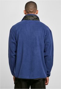 Pullover in pile blu navy con colletto alto e fodera interna nera. Presenta una vestibilità comoda e una superficie liscia, con orli elasticizzati.