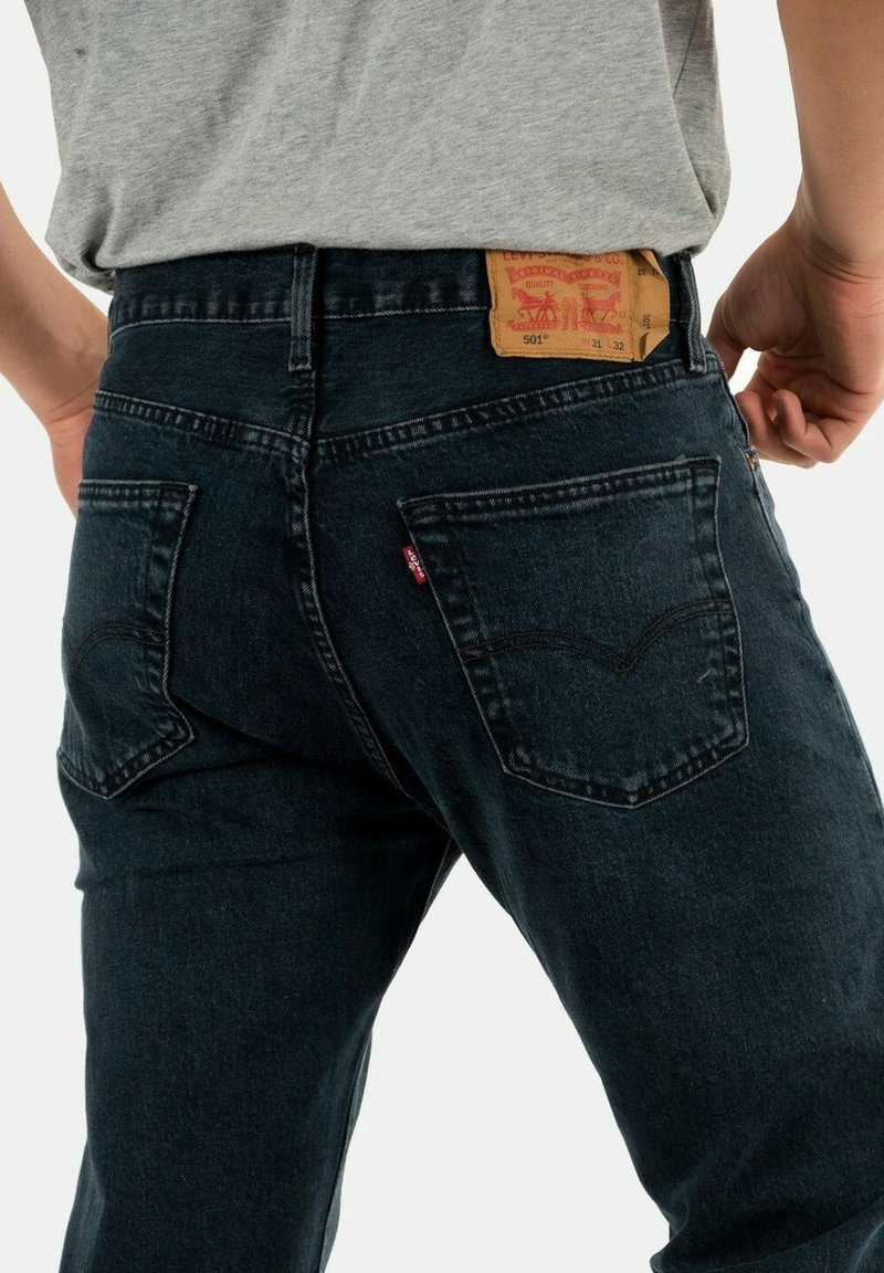 Pantalones vaqueros de mezclilla azul oscuro con un corte recto, que cuentan con dos bolsillos traseros y una etiqueta color marrón en la cintura que muestra los detalles de la marca.