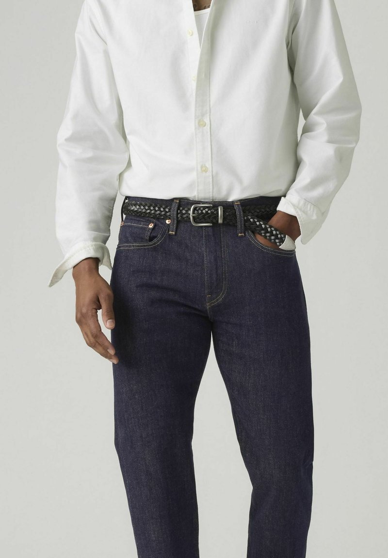 Hombre vistiendo vaqueros azul oscuro con un cinturón negro trenzado y una camisa blanca abotonada, con una mano en el bolsillo y la otra relajada a un lado.