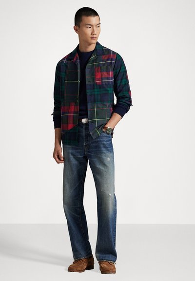 Geruite shirt met marineblauw, groen, rood en paarse tinten; lange mouwen, voorzak. G gedragen met lichtblauwe, verwaterde jeans en bruine schoenen.
