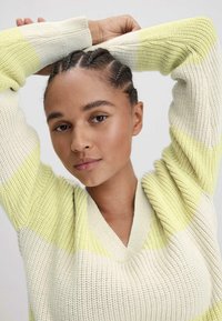 Gestreifte Strickjacke in blassgelb und Weiß, mit V-Ausschnitt und Rippstruktur. Das Model hat geflochtene Haare und die Arme über dem Kopf erhoben.