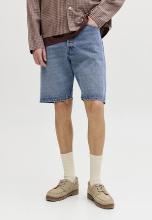 Homme portant un short en denim bleu, des chaussettes beiges, des chaussures beiges à lacets et une chemise marron à carreaux à manches longues, main dans la poche.