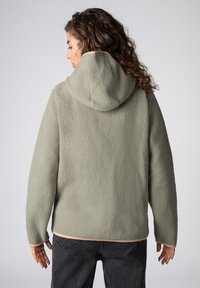 Vrouw met krullend haar, gekleed in een lichtgroene fleece hoodie en donkere spijkerbroek, van achteren gezien tegen een effen achtergrond.