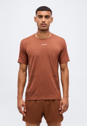 Mand iført brun Salomon sport T-shirt og matchende shorts, stående mod en ensfarvet lys baggrund, vendt fremad med neutralt udtryk.