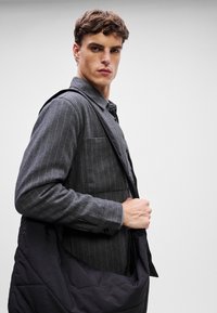 Szürke csíkozott ing gombos gallérral és fekete steppelt táskával, amely a vállon van átvetve. Tartalmaz egy mellzsebet és illeszkedő ujjakat.