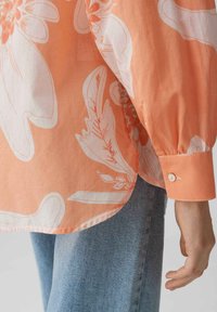 Blusa floreale arancione realizzata in tessuto leggero con motivi floreali bianchi, caratterizzata da polsini con bottoni, vestibilità rilassata e orlo curvo.