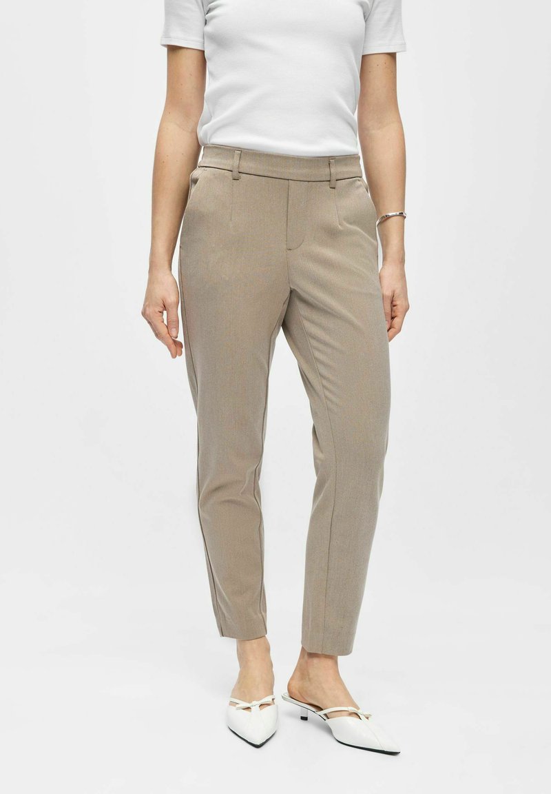 Frau trägt beige, maßgeschneiderte, knöchellange Hosen, weißes Kurzarmtop, silbernes Armband und weiße spitz zulaufende Mules mit niedrigem Absatz.