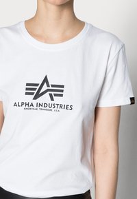 Balta medvilninė marškinėliai su apvaliu kaklu, antkyru juodu Alpha Industries logotipu ir grafiniu dizainu priekyje, trumpomis rankovėmis.