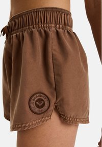 Braune Badeshorts mit elastischem Bund und Kordelzug. Enthält ein rundes Logoschild an der unteren Seite und Seitenschlitze für Flexibilität.