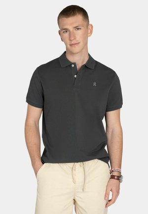 Joven con una camisa polo negra de manga corta y pantalones beige con cordón, llevando un reloj y pulseras en la muñeca izquierda.