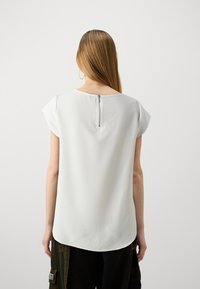 Top bianco a maniche corte con scollo rotondo e dettaglio con zip sul retro, abbinato a pantaloni verde scuro con tasca a toppa.