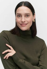 Olivgrön, åtsittande polotröja med ribbade manschetter. Guldörhängen i form av hoops framhäver ansiktet. Slät textur, hög krage.