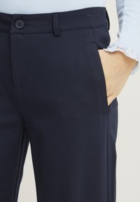 Marinblå byxor med slät front, enkel knappstängning och sidofickor. Tyget ser glatt ut med en lätt struktur och skräddarsydd passform.