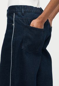 Σκούρο μπλε τζιν με λευκή κάθετη πλευρική ρίγα, ελαστική μέση και χέρι τοποθετημένο στην πίσω τσέπη.