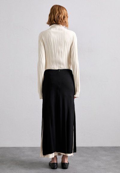 Black skirt with side slits, paired with a crinkled cream blouse. Features long sleeves, a zip closure at the back, and subtle texture variations.