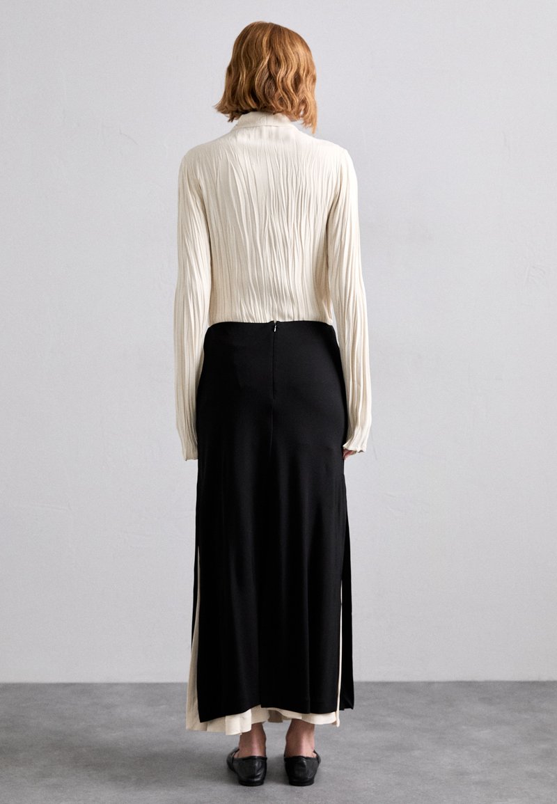Black skirt with side slits, paired with a crinkled cream blouse. Features long sleeves, a zip closure at the back, and subtle texture variations.