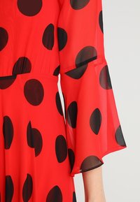 Robe rouge à pois noirs, avec des manches transparentes et un tissu texturé. Le design comprend un buste ajusté et des extrémités des manches évasées.