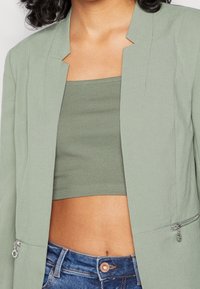 Blazer ajusté vert clair au design épuré, à l'avant ouvert et avec des poches zippées, porté sur un crop top côtelé assorti.