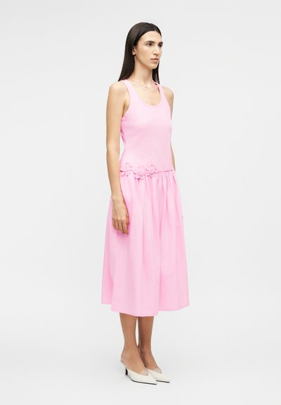 Ženska z dolgimi temnimi lasmi, oblečena v roza obleko brez rokavov z rožnatimi detajli okoli pasu in belimi nizkimi čevlji z ostro konico.
